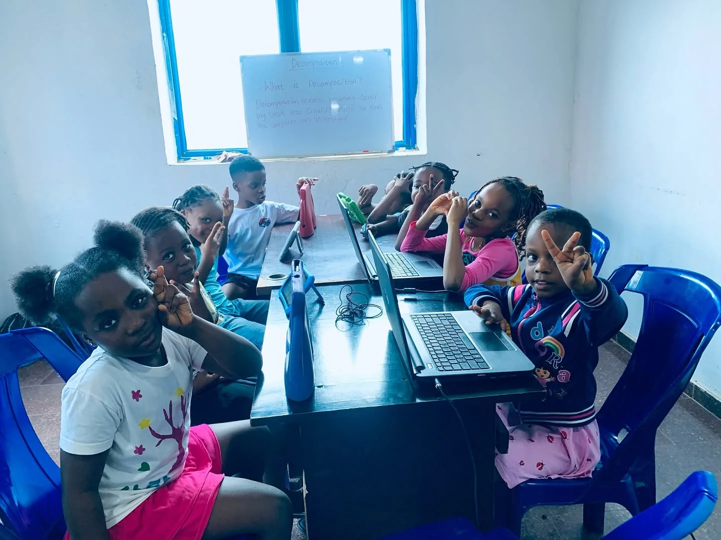 Young MejorTech learners smiling at a shared table of laptops, with a whiteboard lesson on decomposition behind them